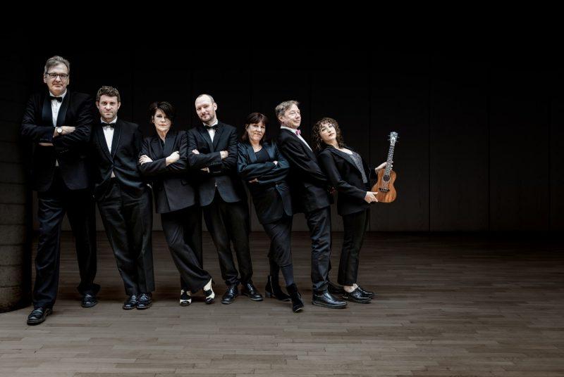 Members of the Ukulele Orchestra of Great Britain, three white women and four white men, stand in a line wearing all black. The women are order from shortest to tallest, left to right, and the men are ordered the same way., and they all lean right towards the tallest man. The woman on the far left holds a ukulele.